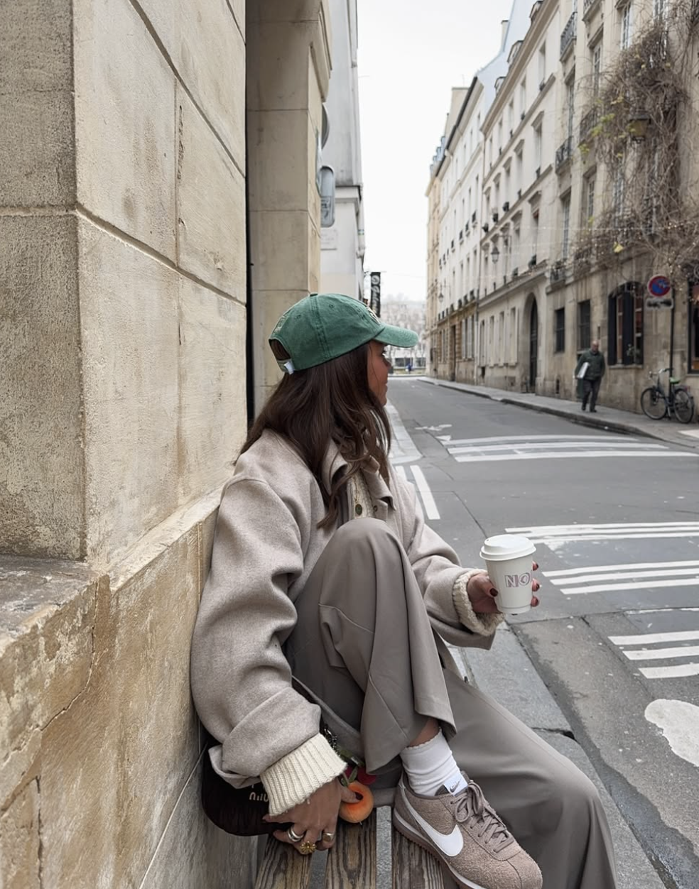 A woman wearing a casual monochromatic grey outfit with a textured jacket and trousers, styled with a green baseball cap and brown Nike sneakers.