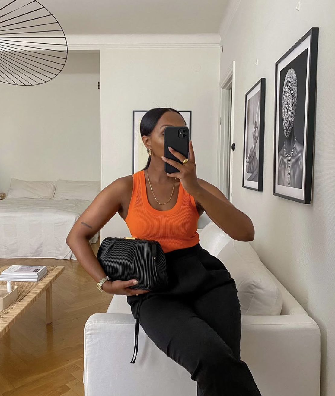 A woman taking a mirror selfie wearing a vibrant ribbed orange knit tank top tucked into high-waisted black trousers, holding a structured black leather clutch bag.