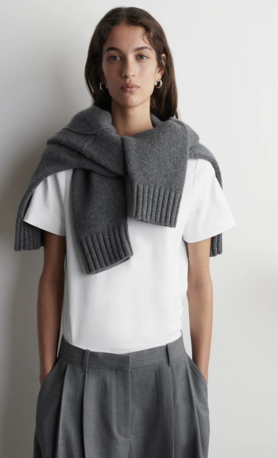 A close-up view of a woman wearing a minimalist white crewneck t-shirt with a medium grey ribbed knit sweater draped over her shoulders like a shawl.