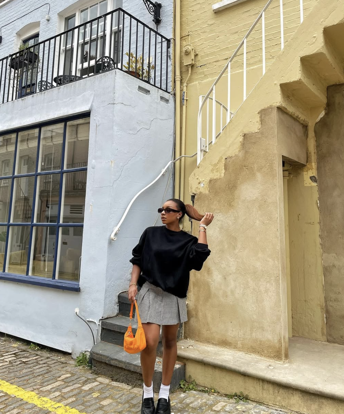 A woman standing on a cobblestone street wearing a black long-sleeve top, a grey pleated mini skirt, white crew socks, and black leather loafers, accessorized with an orange handbag.