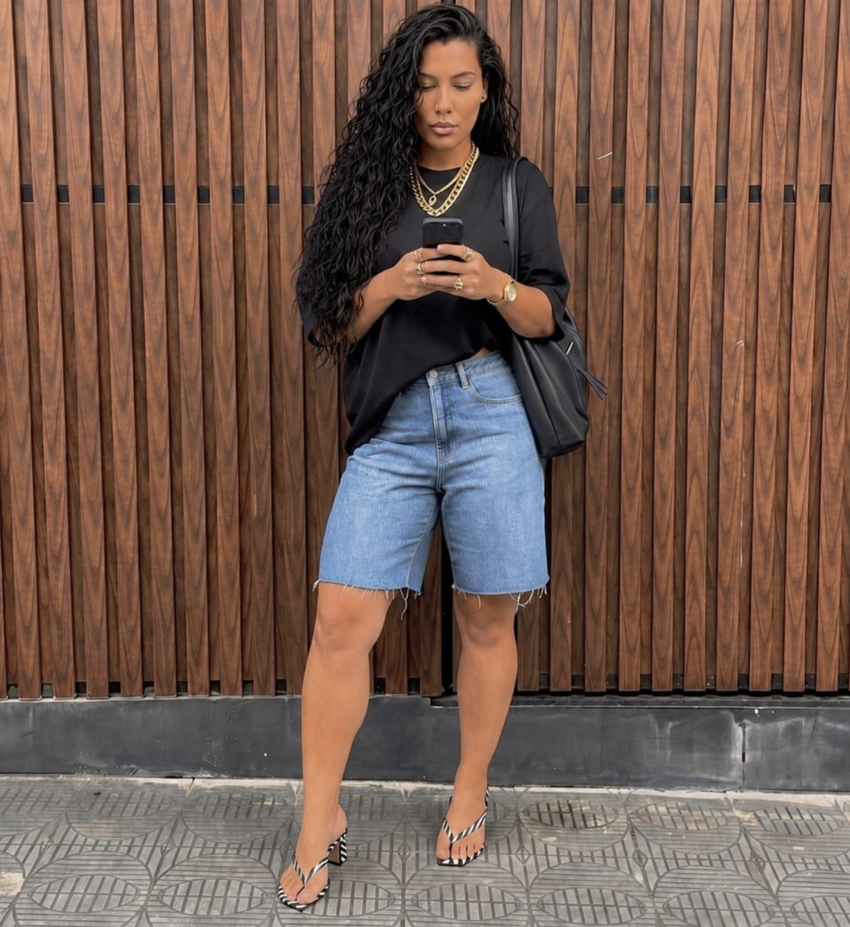 A woman styling high-waisted, light-wash denim Bermuda shorts featuring a raw hem, paired with black kitten-heel thong sandals.