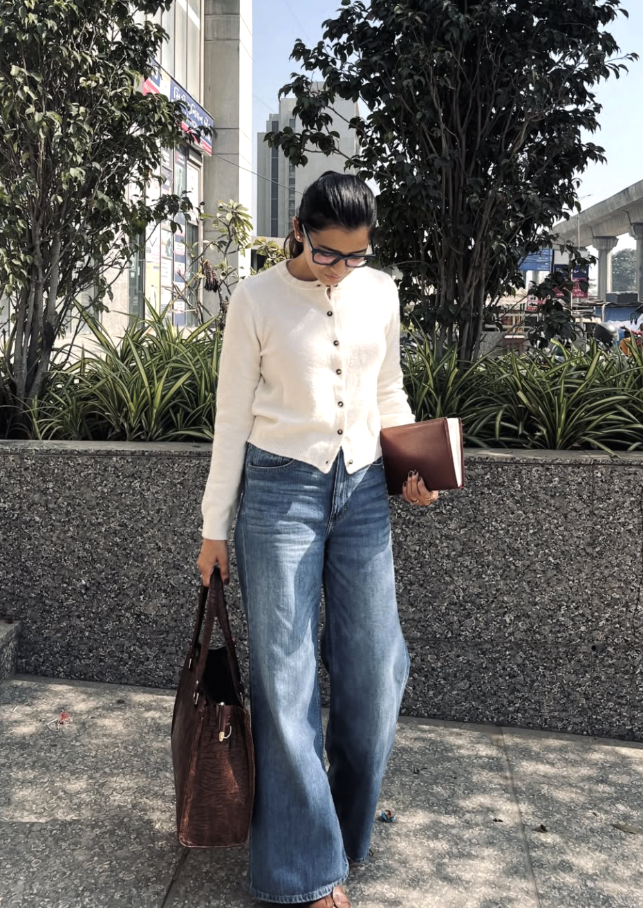 A woman wearing baggy medium-wash jeans with a white button-up cropped cardigan and a large brown leather tote bag to demonstrate a balanced waistline silhouette