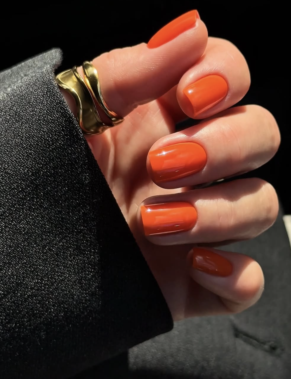 Close-up of a hand with short, square-shaped nails showcasing a vibrant, high-gloss tangerine orange manicure, styled with a bold, wavy chunky gold statement ring and a black blazer sleeve.