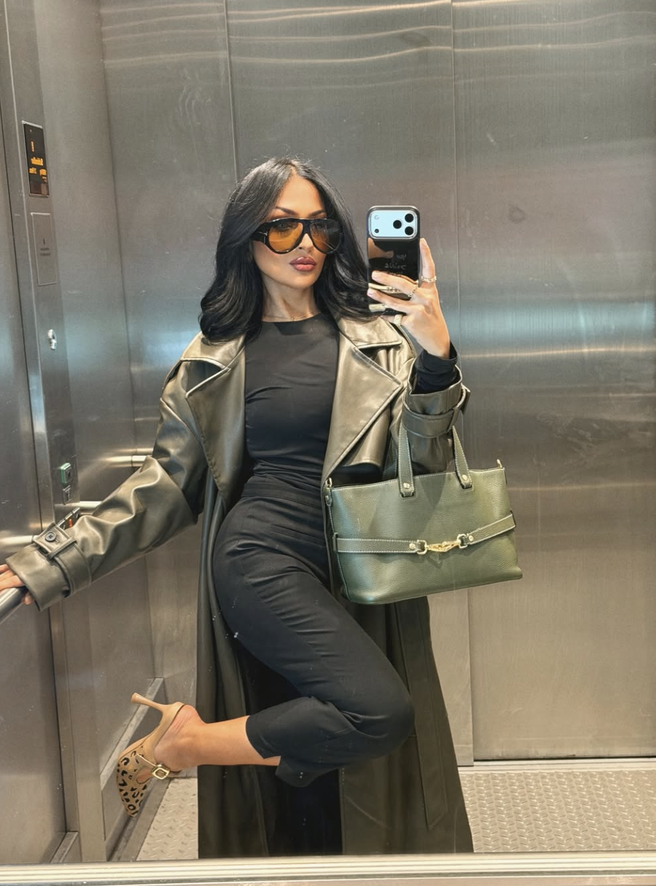 A fashion creator taking a mirror selfie wearing black high-waisted capri pants, a black bodysuit, a long olive green leather trench coat, and leopard print pointed-toe slingback heels.