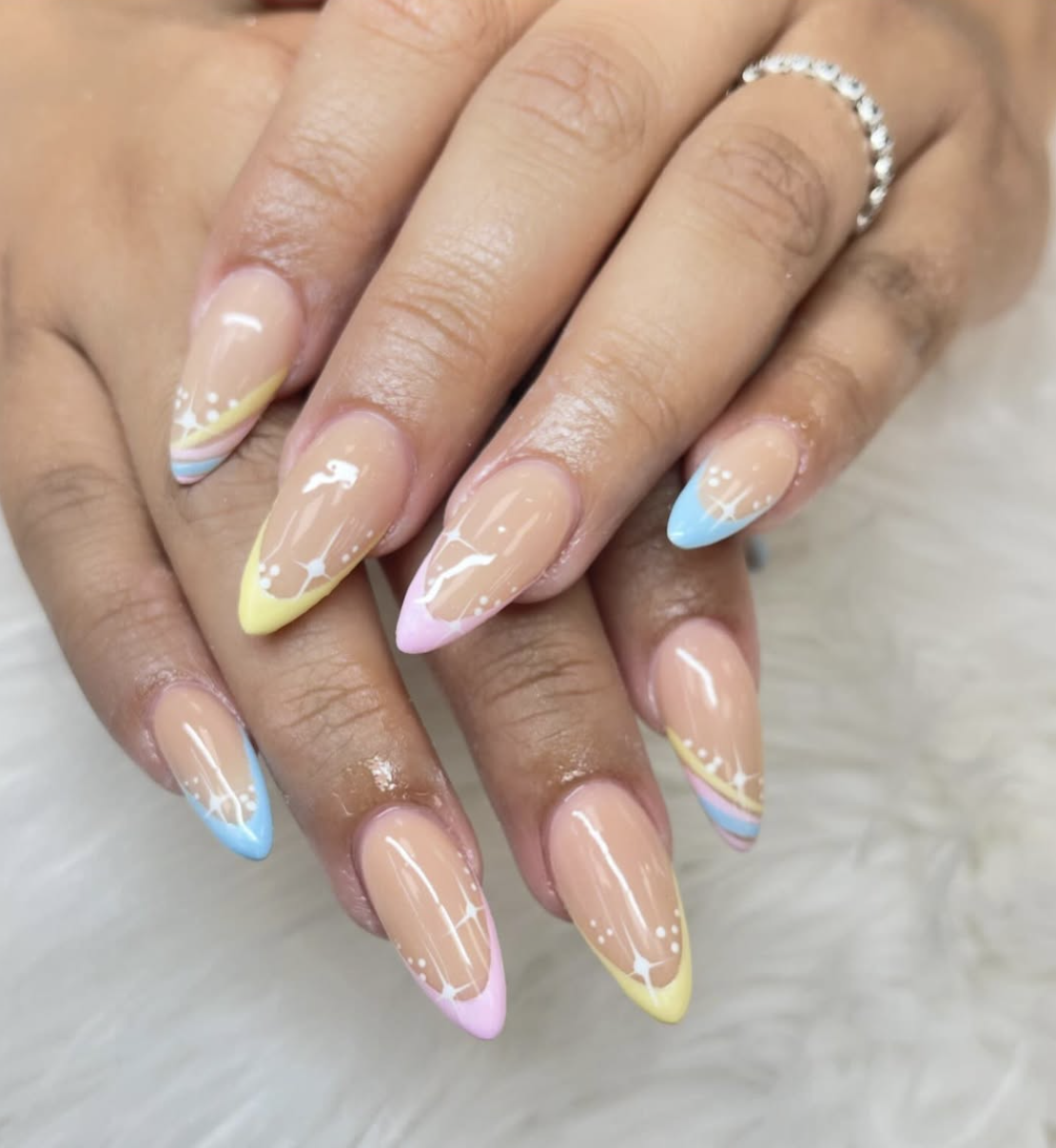 A close-up of a hand with almond-shaped acrylic nails featuring a sheer nude base and mismatched pastel French tips in yellow, blue, and pink, decorated with delicate white hand-painted