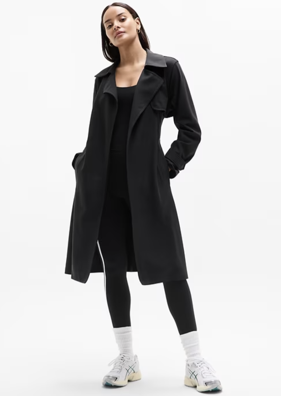 A woman wearing the black Athleta Stellar Trench coat, a lightweight and water-resistant performance jacket with a classic lapel and a streamlined, modern fit.