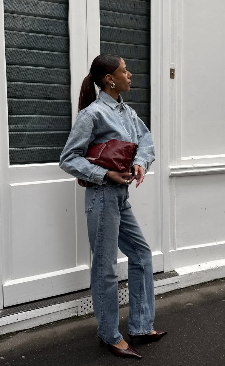 Side profile of a woman wearing a light wash denim shirt, cigarette jeans, and carrying a burgundy leather clutch.