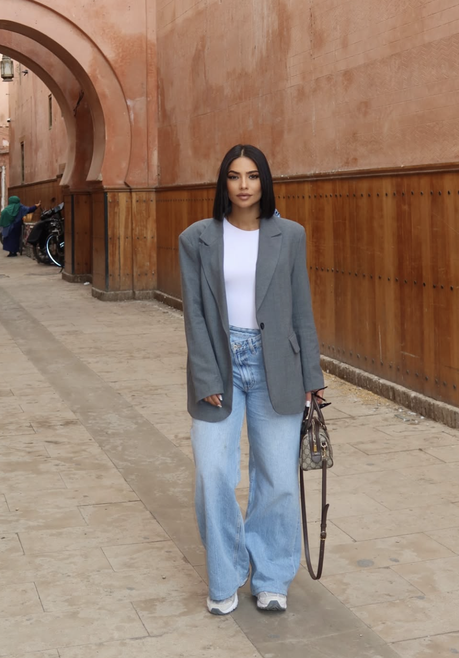A street style lookbook image of a woman wearing a crisp white crew neck t-shirt layered under a grey oversized blazer and paired with light-wash baggy jeans. She is carrying a designer shoulder bag, illustrating a master-of-layers outfit formula.