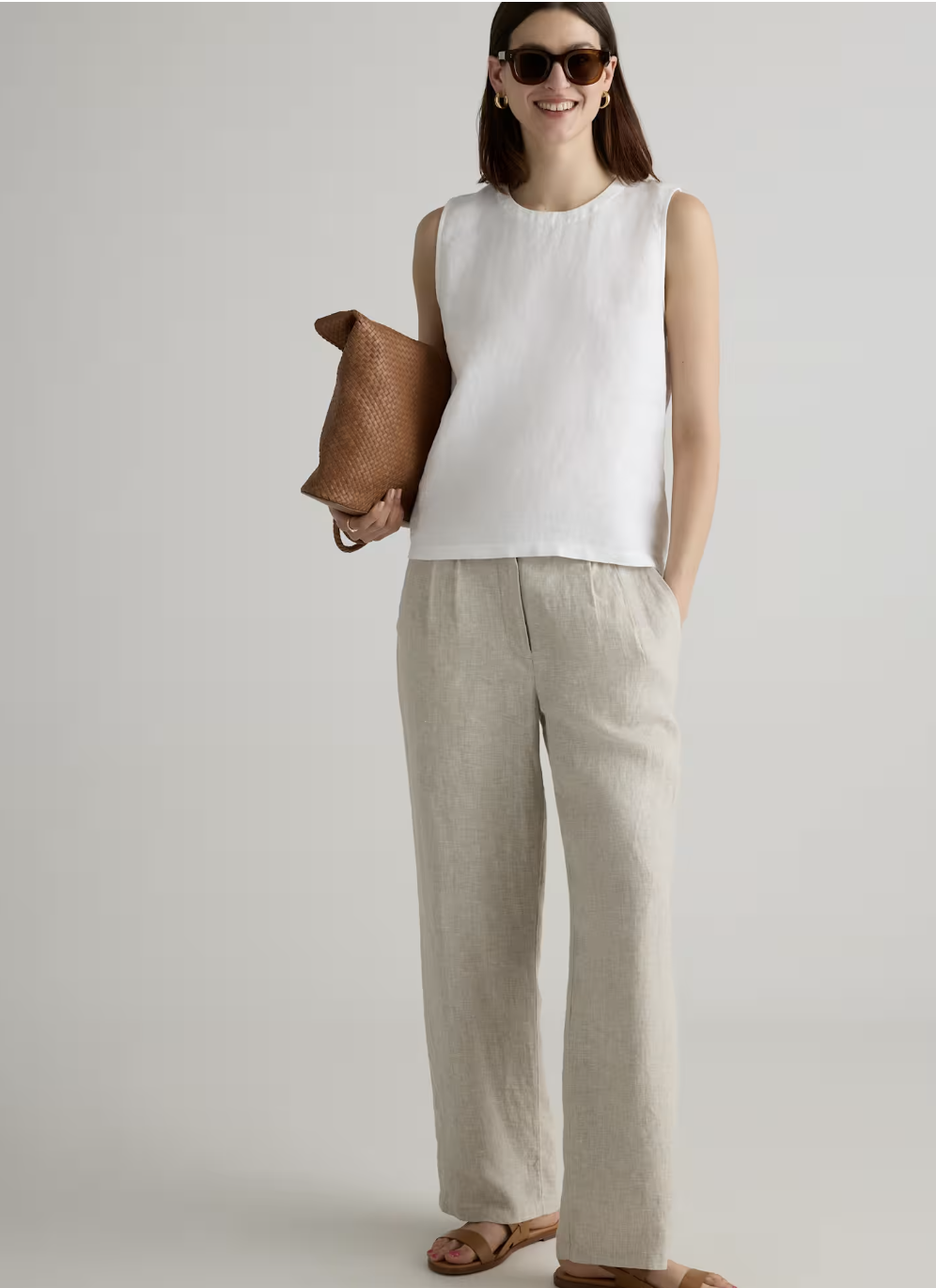 A woman styling a minimalist summer outfit featuring a crisp white sleeveless top tucked into pleated, wide-leg tan linen trousers, paired with flat brown leather sandals.