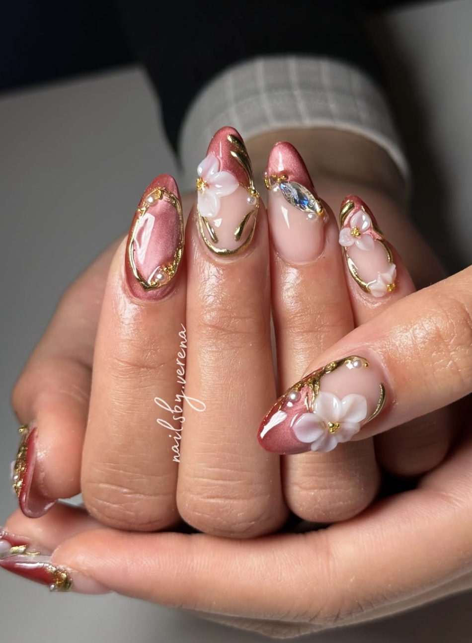 Long almond-shaped nails featuring a mix of metallic rose gold polish, 3D white flower petals with pearl centers, and intricate gold wire detailing with embedded crystals.