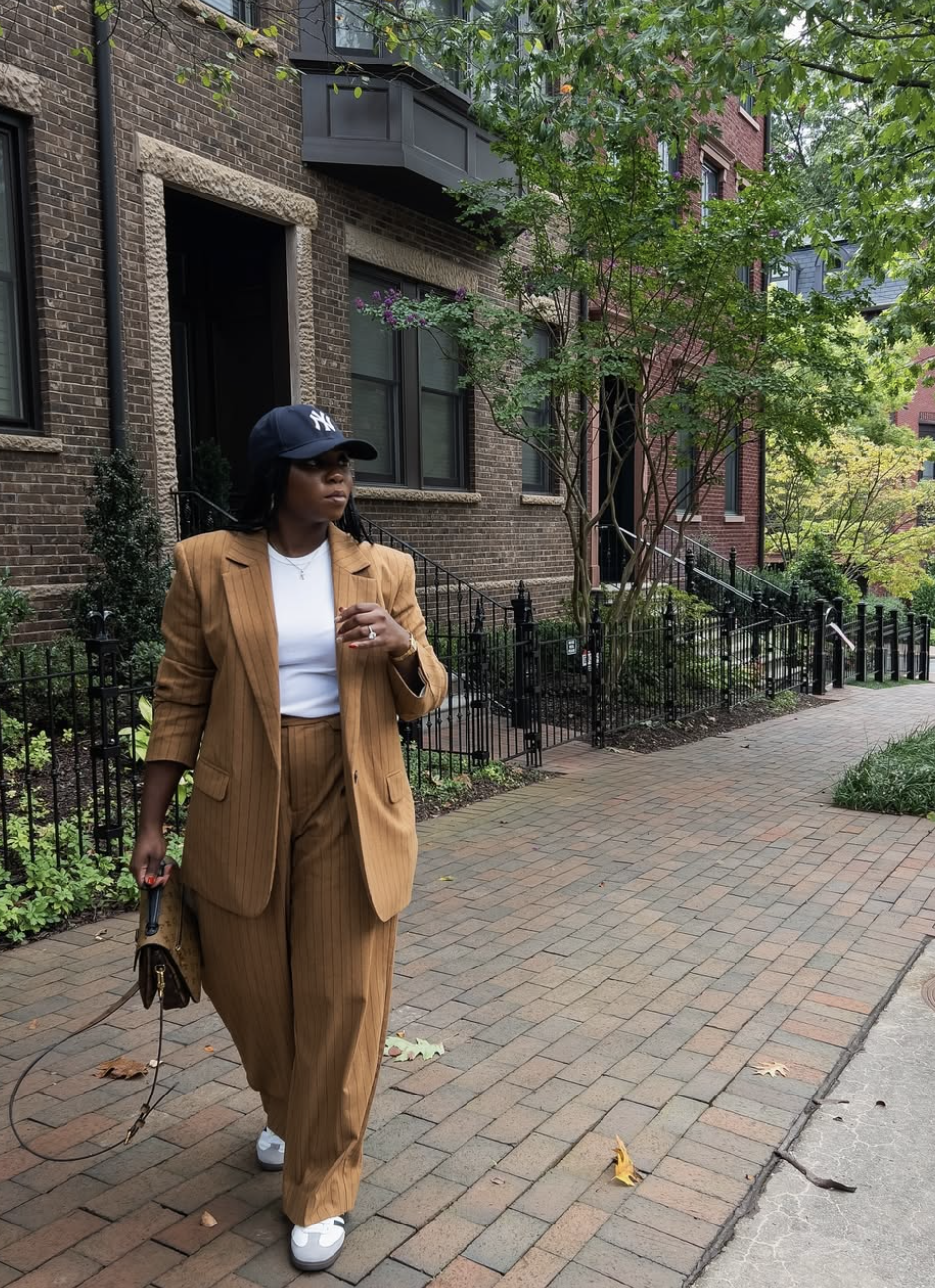 A street style lookbook image of a woman wearing a white t-shirt layered under a tan oversized blazer and paired with matching trousers. She is wearng a black cap and walking down a city sidewalk, illustrating how to layer basics