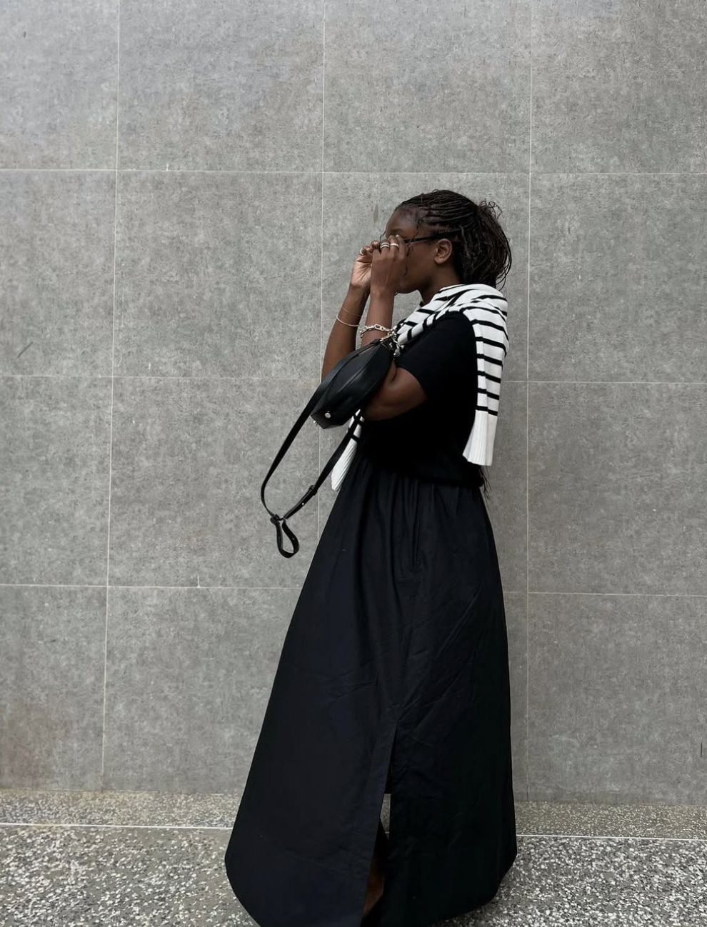 A woman from the side wearing a black maxi skirt with a high slit, styled with a black top and a black and white striped sweater slung over her shoulder.