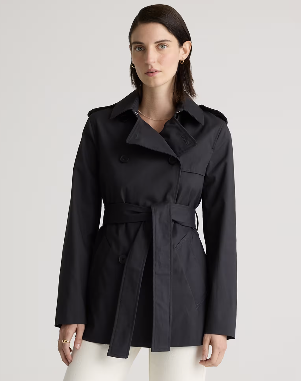 "A woman wearing a black Quince Comfort Stretch Short Trench Coat with a cinched belted waist, structured collar, and double-breasted buttons."