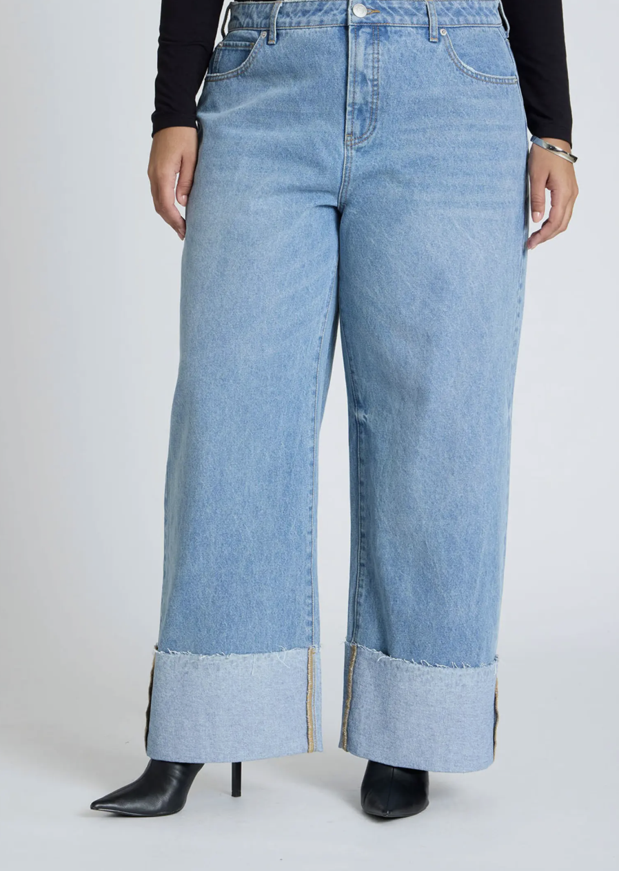 A close-up of light blue wide-leg jeans featuring a dramatic tall folded cuff at the hem, styled with black pointed-toe stiletto boots.