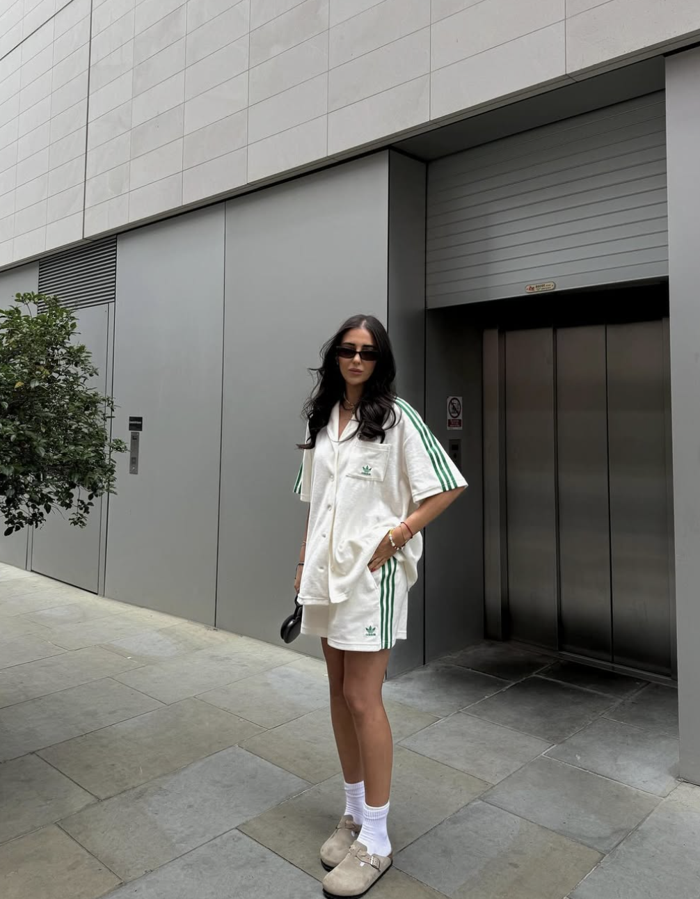 A woman standing on a city street wearing a monochromatic summer outfit featuring an oversized cream button-down shirt paired with matching white track shorts, white crew socks, and white sneakers.