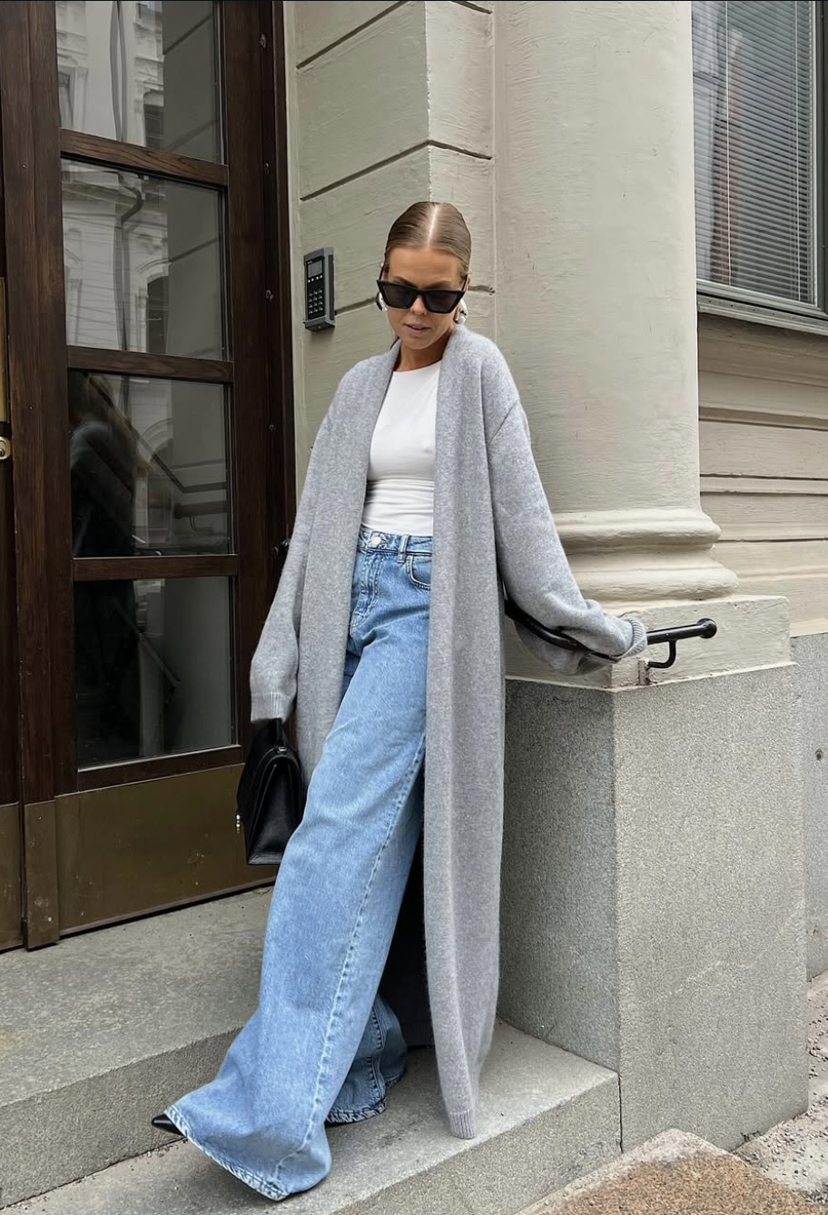 A woman wearing light-wash wide-leg jeans, a white top, a long floor-length gray cardigan, sunglasses, and a black shoulder bag.