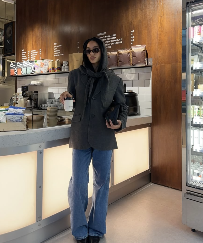 A woman wearing an oversized grey blazer layered over a dark grey hoodie and a matching knit scarf, holding a coffee cup for a cozy street-style look.