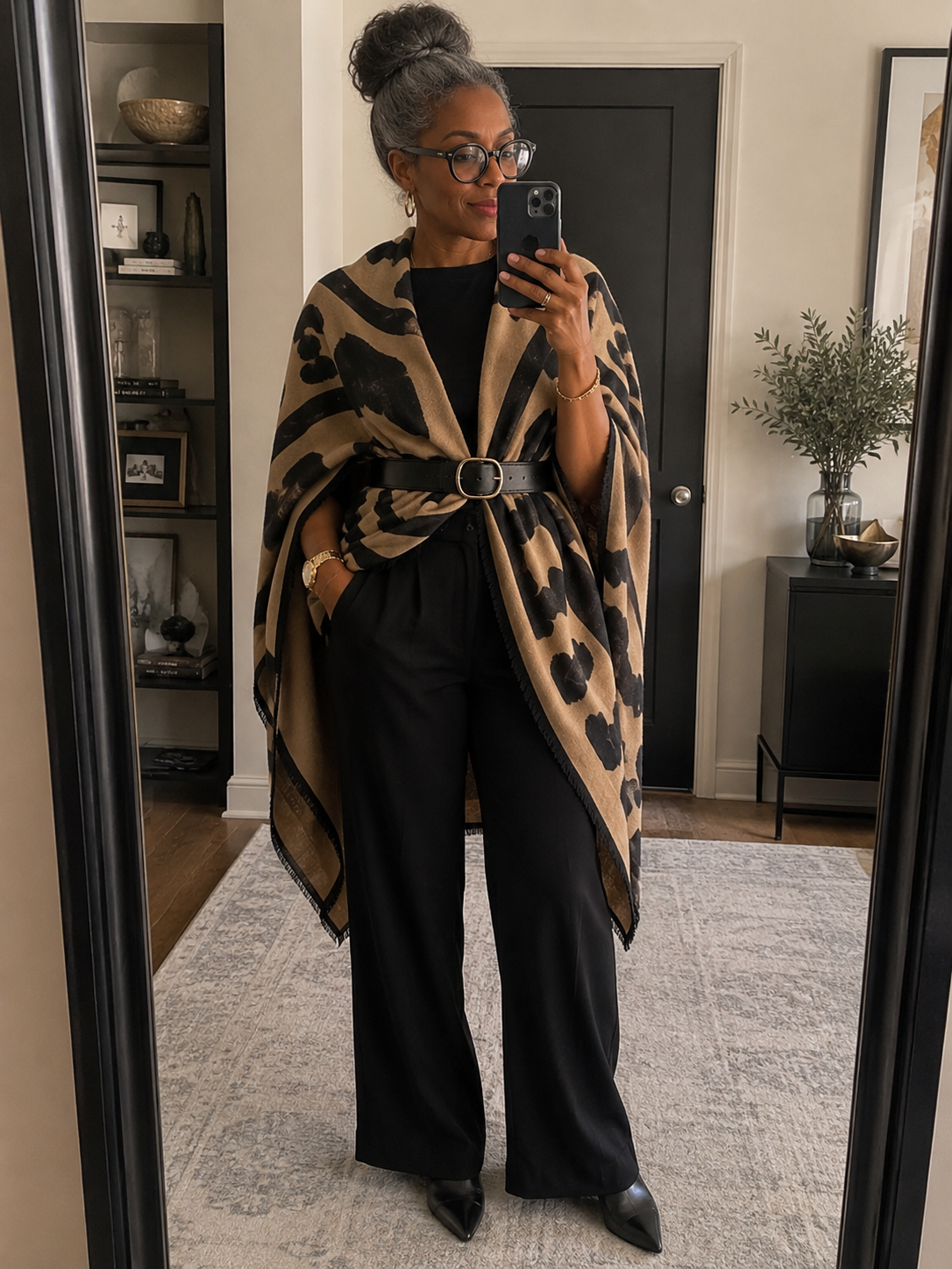 A close-up shot of a woman with her hair in a high bun, wearing a large neutral and black leopard print scarf styled as a belted cape over an all-black outfit.