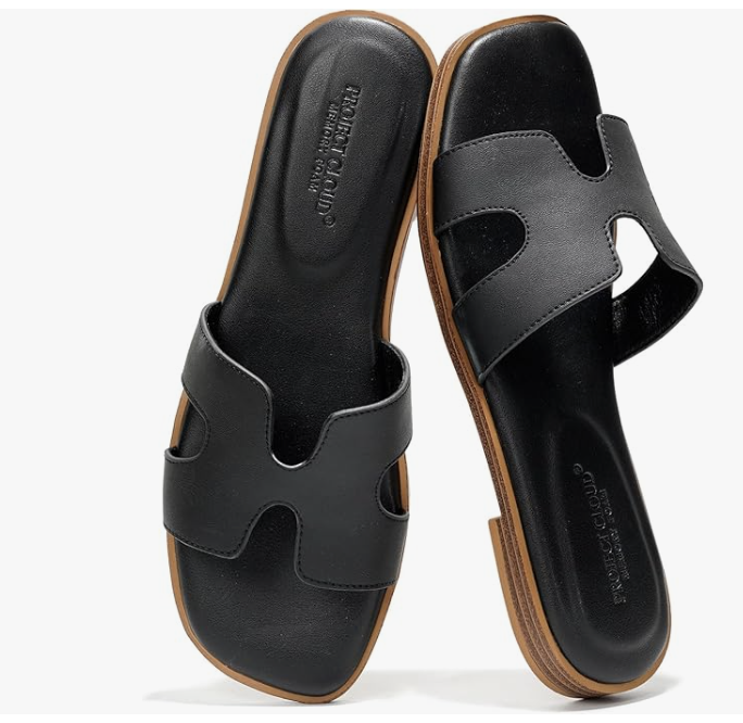 A close-up of a pair of classic black leather flat slide sandals with a distinctive cutout design and a comfortable tan sole.====
