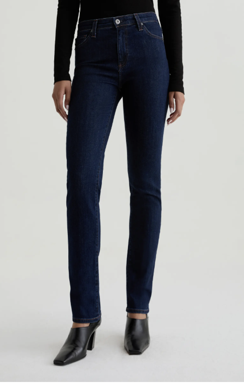 A close-up of a person wearing AG Mari Extended Slim Straight Leg Jeans in a dark-wash denim, styled with a black long-sleeve top and black boots.