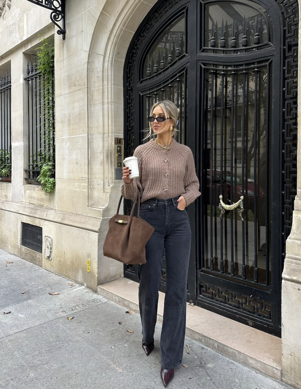 Fashion influencer wearing a chunky taupe cardigan tucked into dark wash bootcut jeans, holding a brown tote bag and coffee cup.