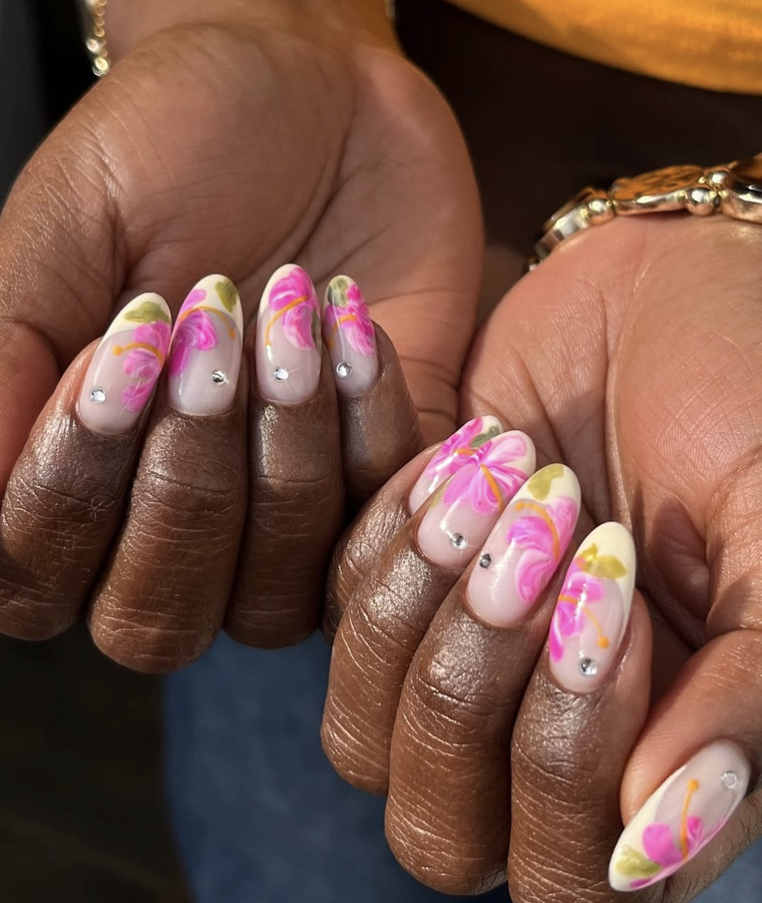 A close-up of almond-shaped nails with a creamy white French manicure, featuring hand-painted pink and orange hibiscus flowers with tiny rhinestone accents on a deep skin tone.