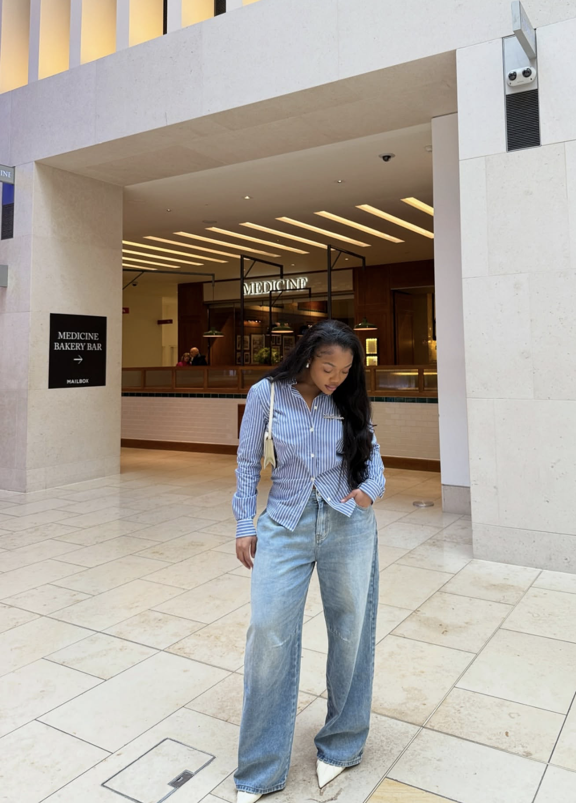 A woman wearing a blue and white striped button-down shirt tucked into oversized light wash baggy jeans with white pointed-toe heels.