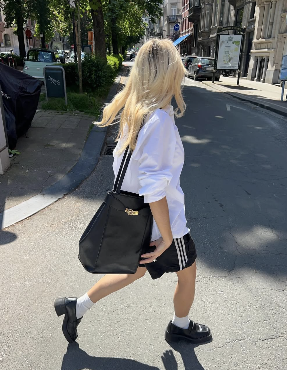 A woman walking down a city sidewalk wearing an oversized white button-down shirt paired with black athletic track shorts, white crew socks, chunky black loafers, and carrying a large black shoulder bag.