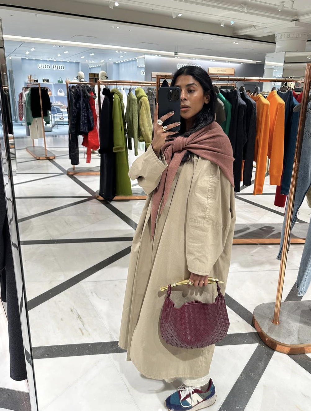 A mirror selfie of a woman in a clothing boutique wearing a mauve-brown silk scarf loosely draped and knotted over her shoulders, paired with an oversized beige trench coat.