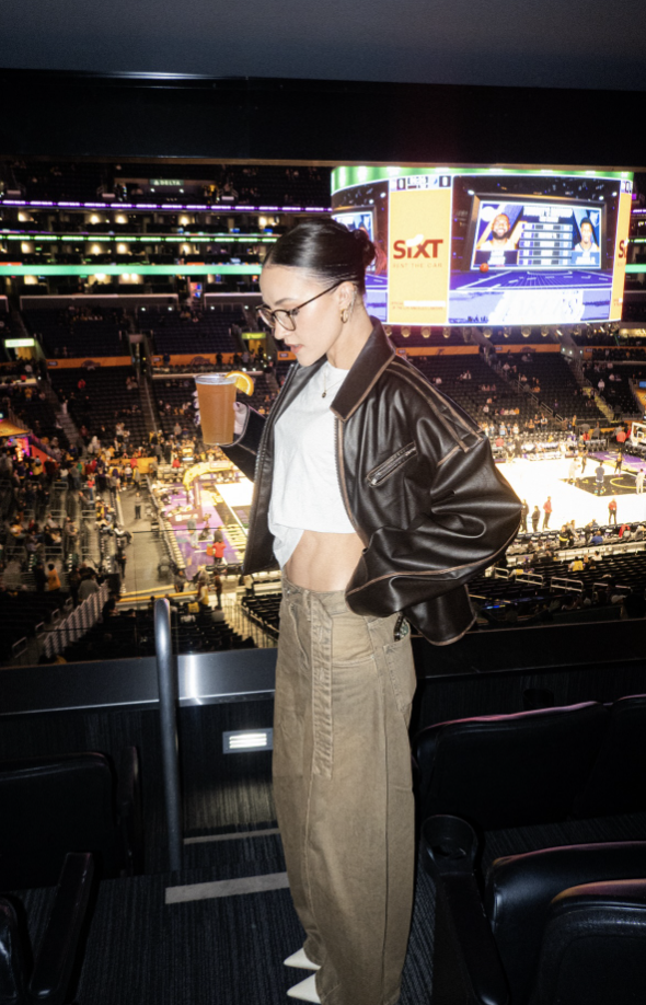 Woman at a basketball game wearing a black leather jacket, white top and taupe wide-leg trousers.