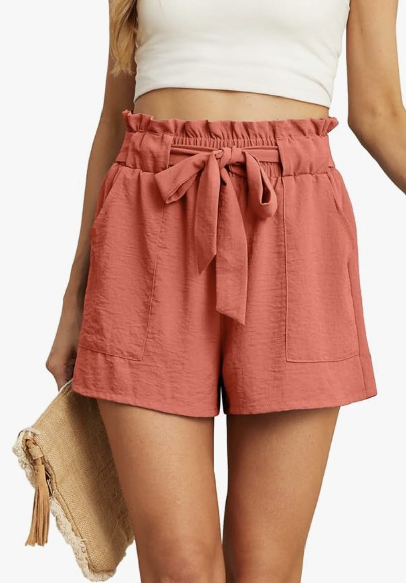 A woman wearing rose-colored high-waisted pleated shorts with a tie-waist bow, designed to add volume and dimension to skinny legs.