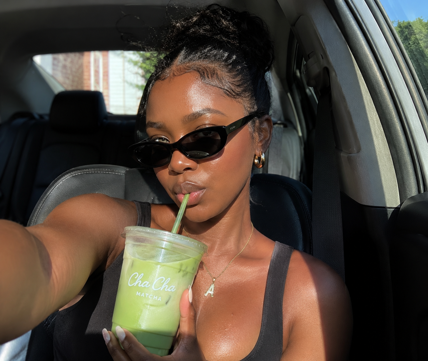 A close-up selfie of a person with dewy, sun-kissed skin and sleek updo wearing dark sunglasses, sitting in a car and sipping an iced matcha latte through a straw.