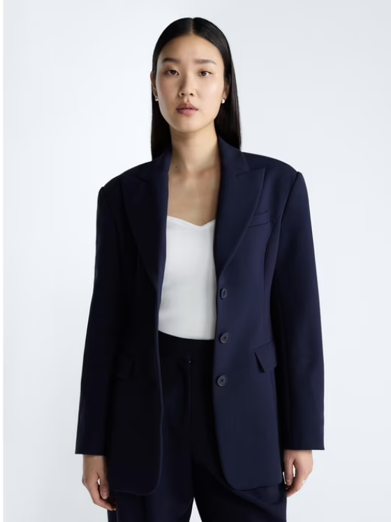 A Black woman wearing a tailored, navy blue single-breasted blazer over a white ribbed top, showcasing a "Miranda Priestly" inspired investment piece from the Devil Wears Prada x Walmart collection.