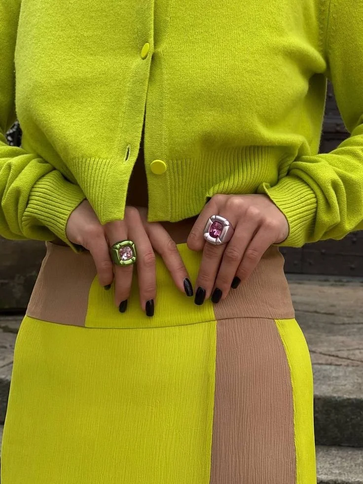 A fashion editor-approved styling for Spring 2026, featuring a vibrant Wasabi green cardigan and matching color-blocked skirt with oversized gemstone rings and black nail polish.