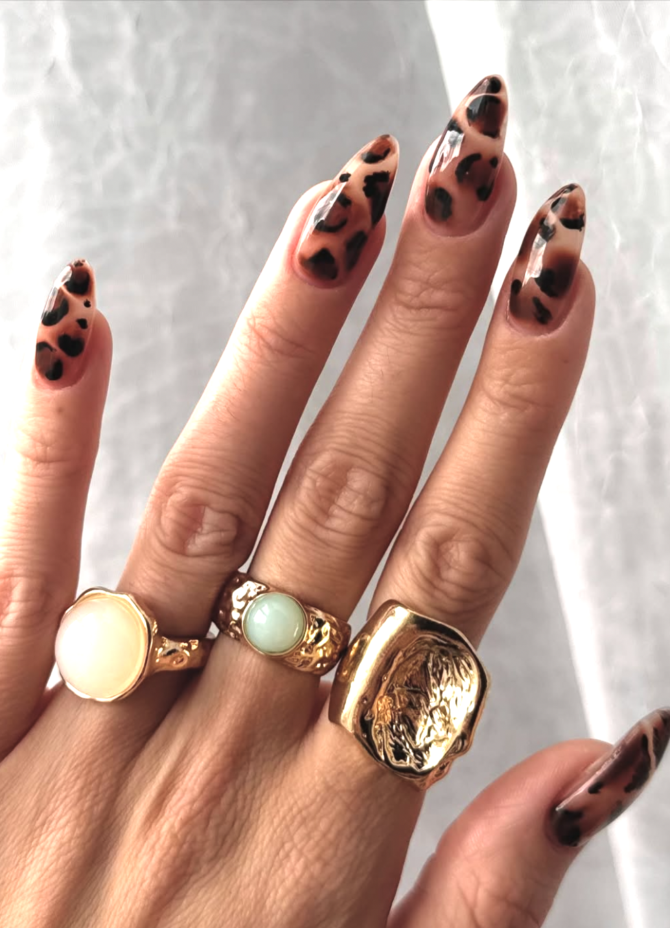 A close-up of hands featuring leopard print nail art on long almond nails, accessorized with chunky vintage gold rings.