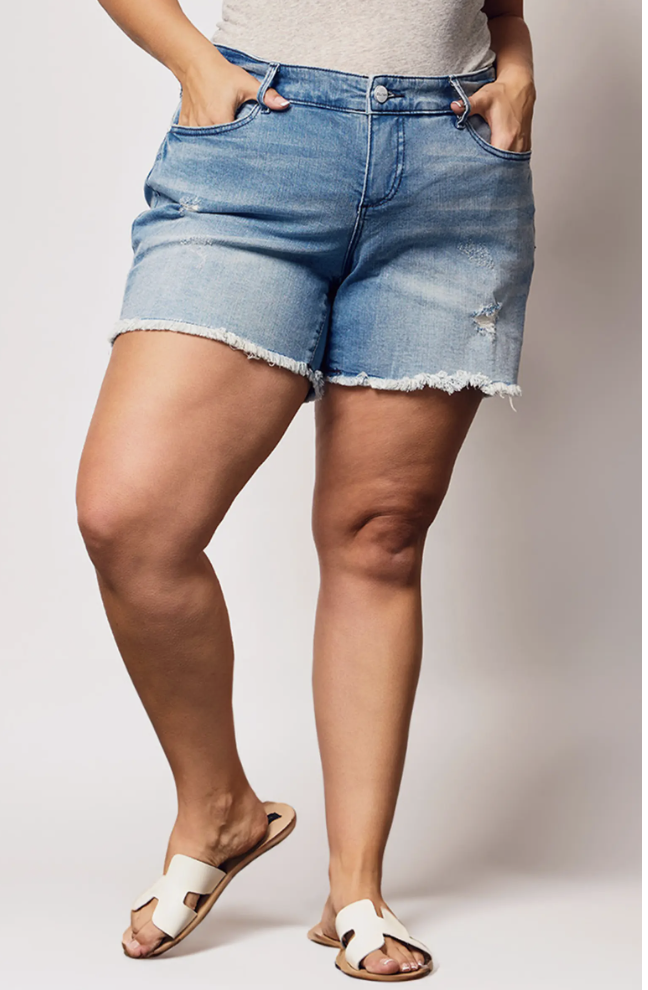 A woman with fuller legs wearing light-wash SLINK Jeans raw hem cutoff denim shorts, featuring a wide A-line leg opening to prevent riding up on thick thighs.