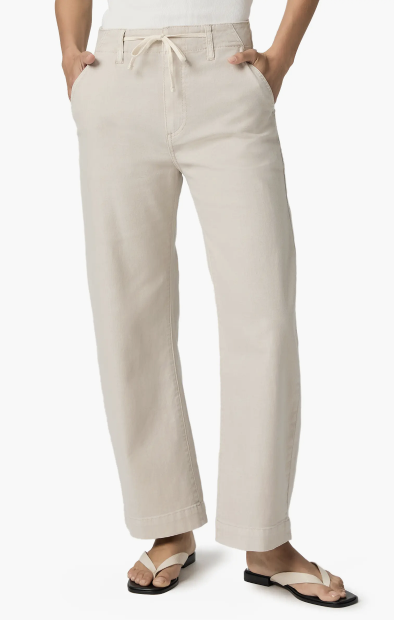 A close-up of a person wearing PAIGE Arellia drawstring high-waist barrel leg jeans in a light sand color, featuring a relaxed trouser-style fit and styled with white thong sandals.