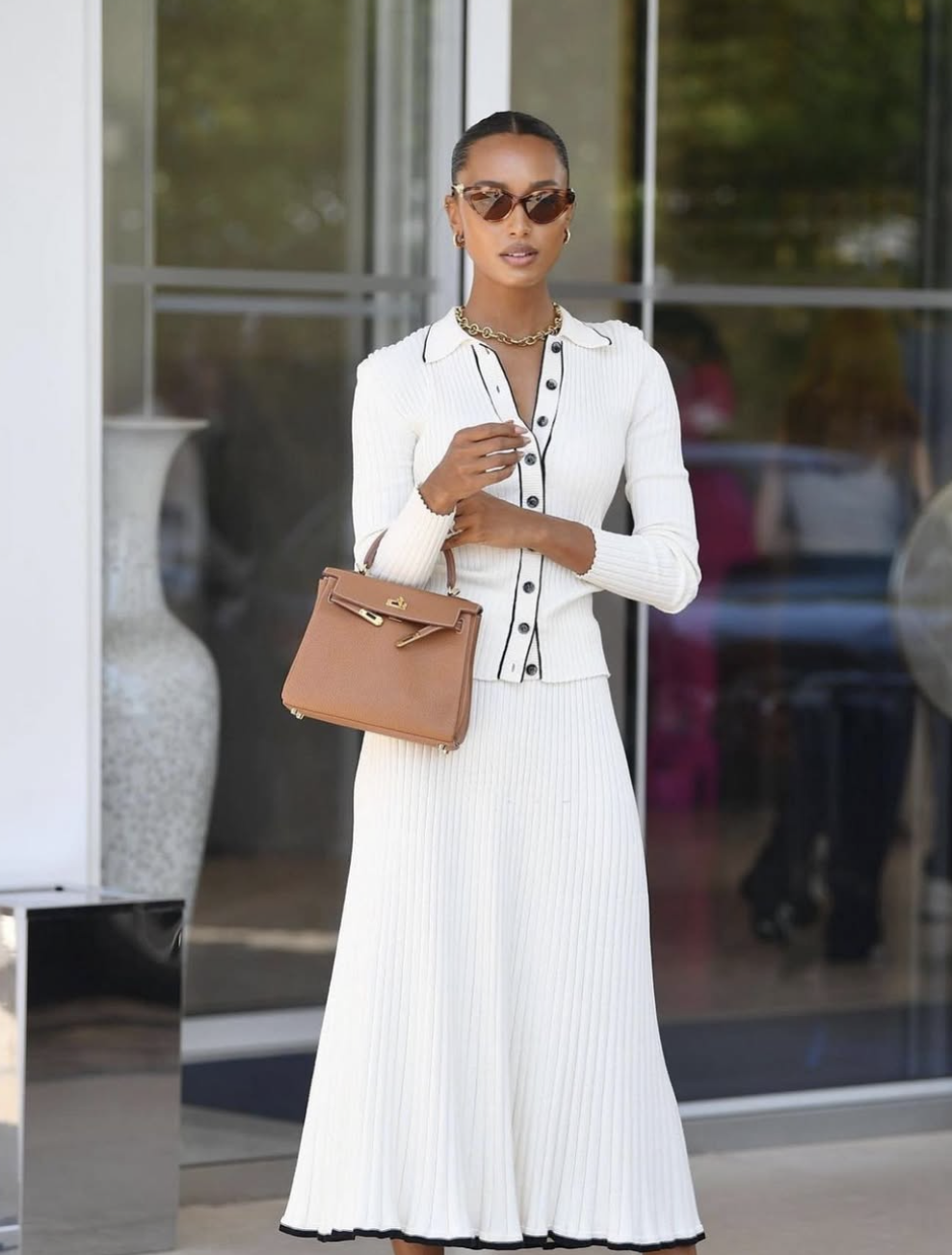 A woman styling a white button-up cardigan as a blouse with a pleated tan midi skirt and a brown leather bag for a professional work outfit.