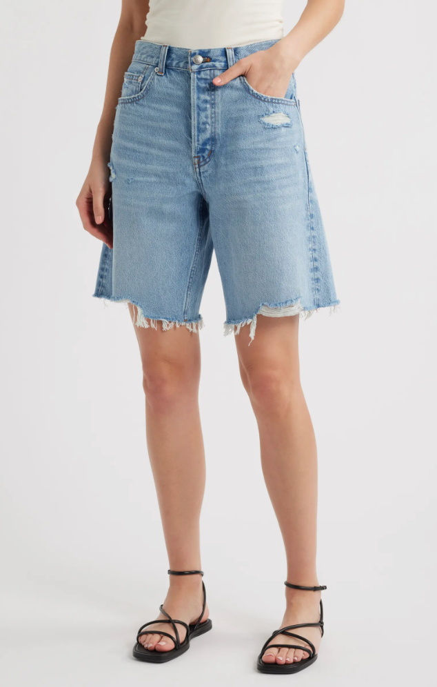 A woman wearing light-wash Madewell Essex raw hem baggy denim Bermuda shorts styled with a white top and black strappy sandals.