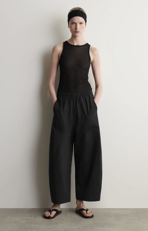 A woman wearing high-waisted black subtle barrel-leg trousers paired with a black sleeveless top and minimalist black sandals.