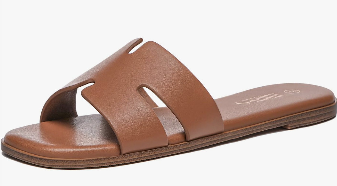 A close-up view of a minimalist tan leather flat slide sandal featuring a wide dual-strap cutout design and a classic open back.