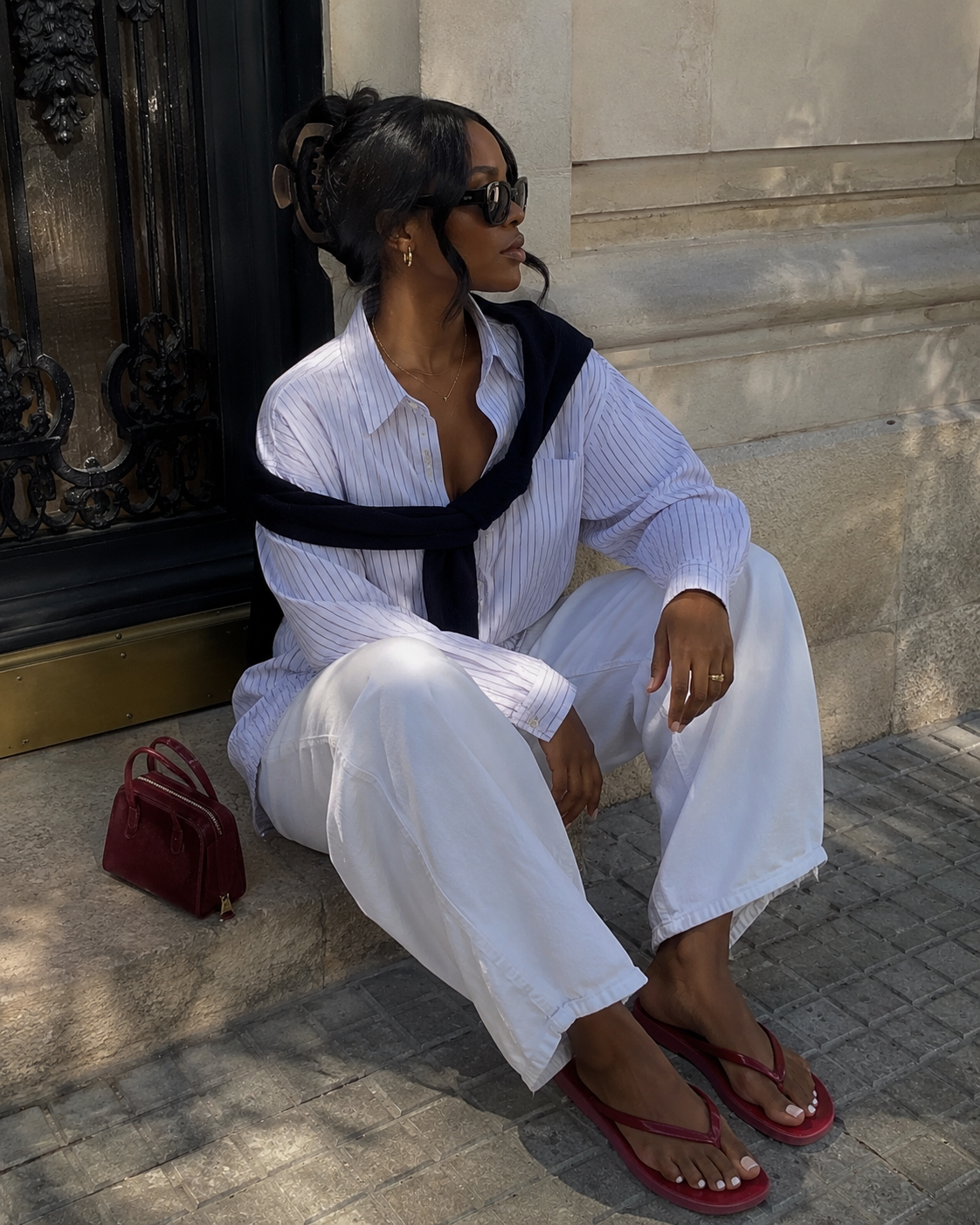 : A person wearing high-waisted white wide-leg trousers, a white button-down shirt with a black sweater draped over the shoulders, and minimalist red leather flip flops.