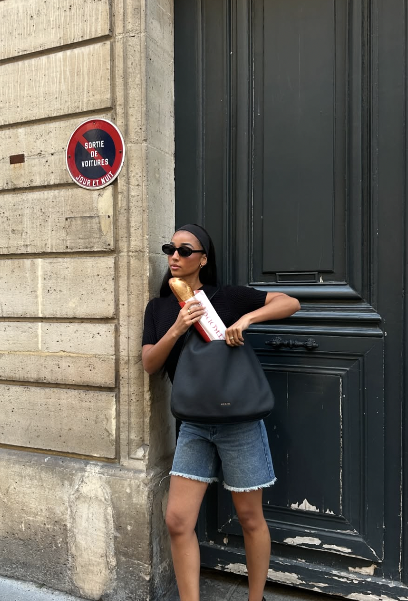 A woman wearing a fitted black short-sleeve top and raw-hem denim shorts styled with a black headband, black sunglasses, and a large black leather shoulder bag holding a baguette.