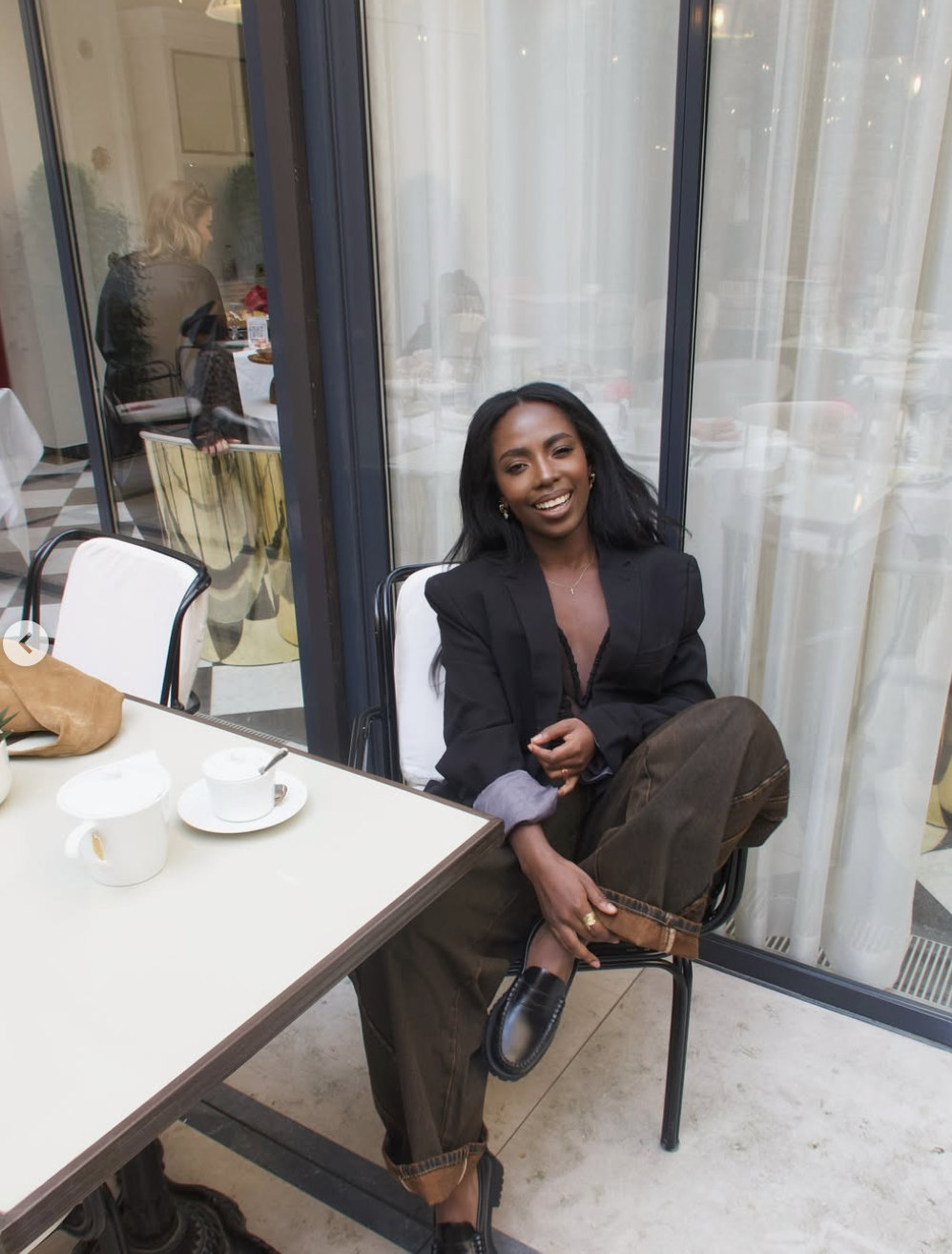 A woman sitting at an outdoor cafe table wearing a tailored black blazer over a black top, paired with chocolate brown wide-leg trousers and black loafers.