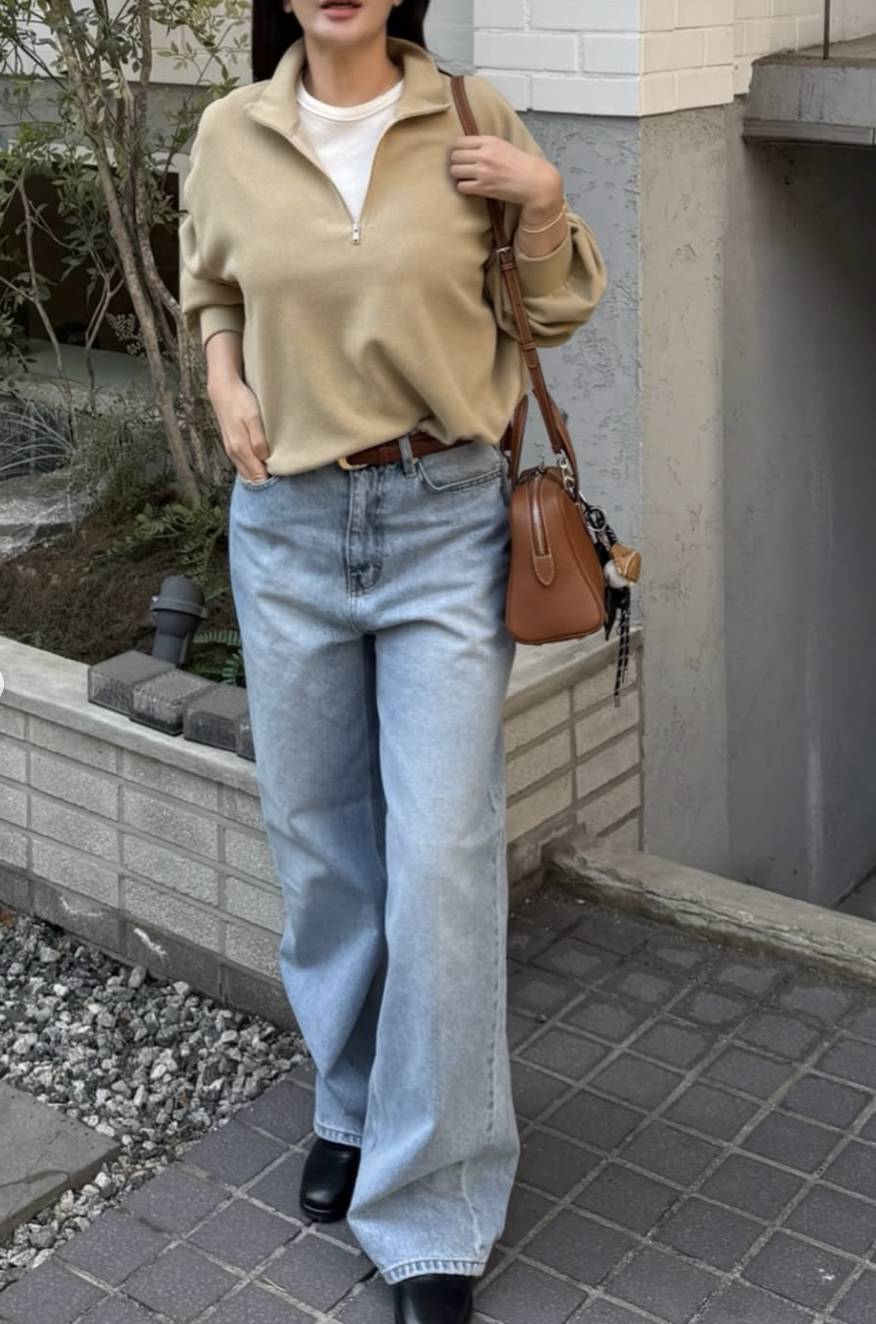 A woman wearing a beige oversized half-zip pullover sweater over a white t-shirt, styled with light wash jeans, a brown leather belt, and a matching brown shoulder bag.