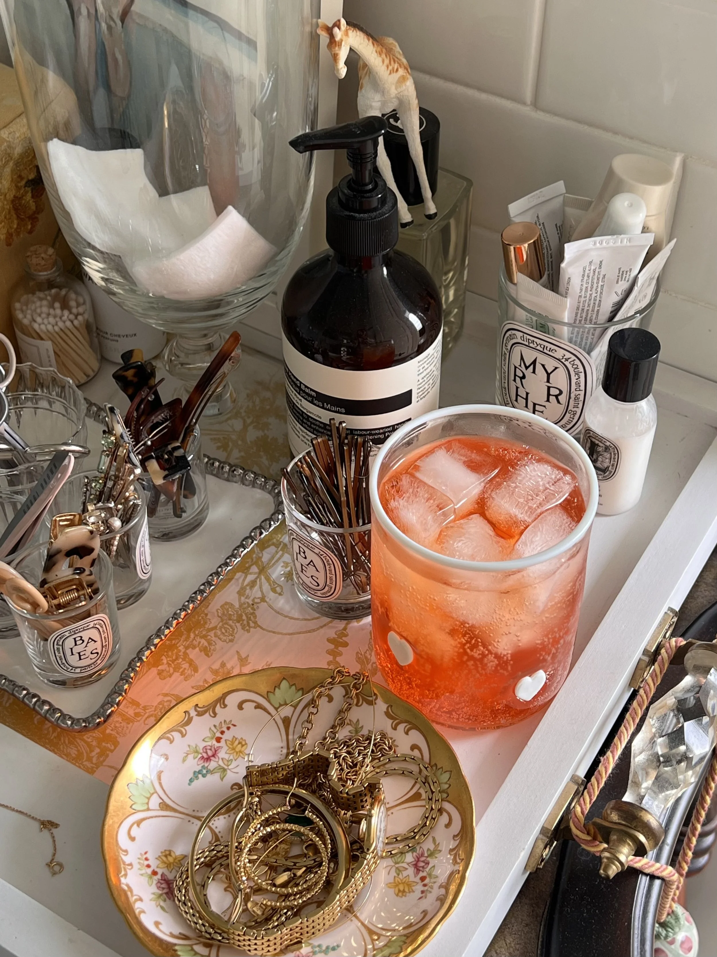 Bathroom vanity tray with a hand soap bottle, makeup brushes, an iced drink and gold jewelry in a floral porcelain dish