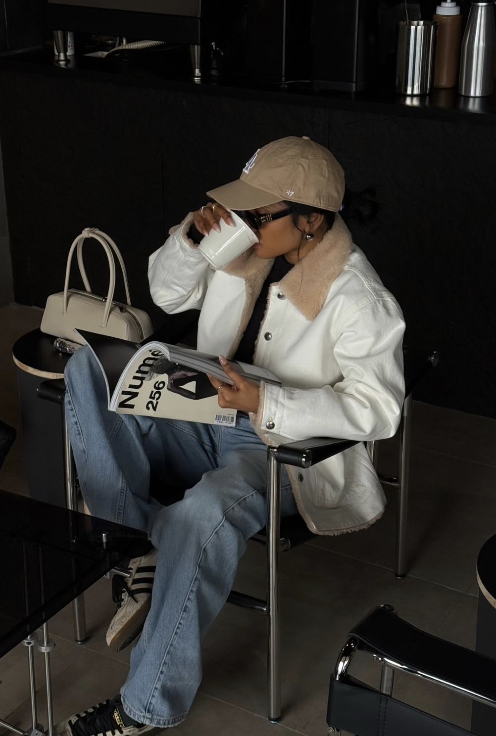 A woman sitting in a cafe wearing light blue jeans, a white bomber jacket with a tan shearling collar, a beige baseball cap, and white sneakers.