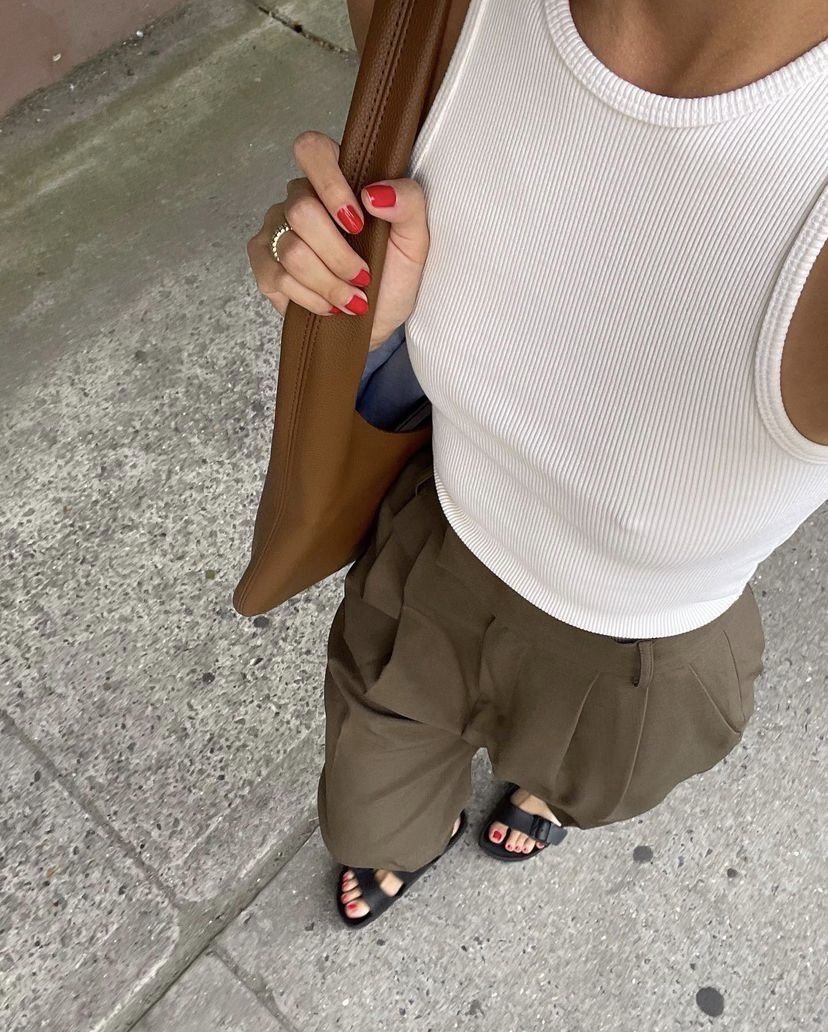 A close-up stylist flatlay of a white ribbed tank top tucked into olive green tailored trousers, accessorized with a brown leather tote bag, black strappy sandals, and red nail polish.