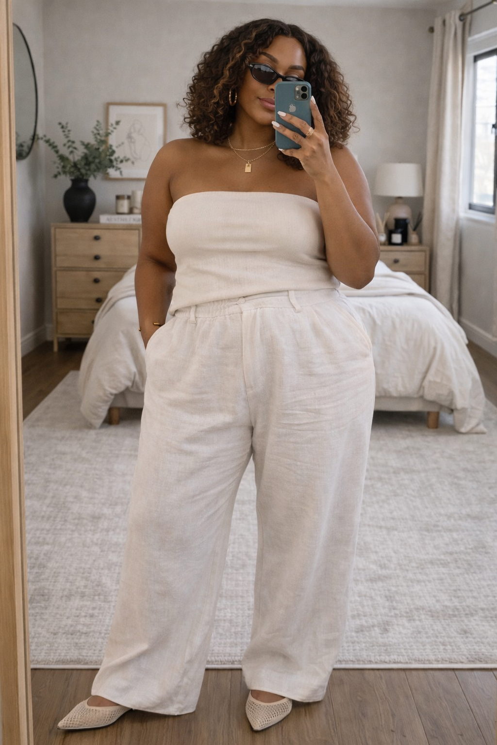 A woman taking a mirror selfie wearing a white strapless tube top and matching high-waisted wide-leg linen pants in a bright bedroom.