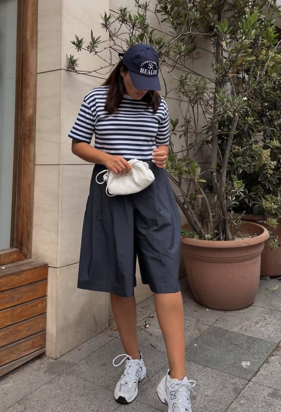 A woman wearing a navy blue and white striped t-shirt, oversized navy Bermuda shorts, a navy baseball cap, and chunky white platform sneakers.
